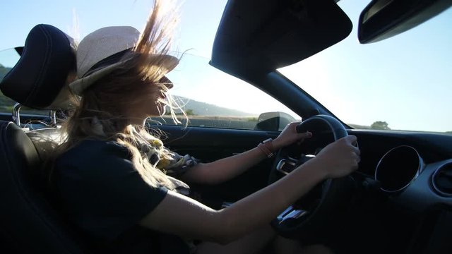 Blond model girl is driving in convertible car in white hat in California having her hair waving in the wind