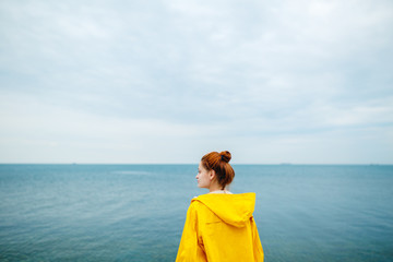Back view of redhead woman in yellow raincoat looking away on background of blue ocean.