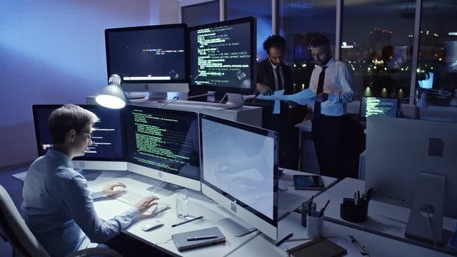 Female programmer typing computer code on computer workstation in the office at night while two coworkers discussing documents