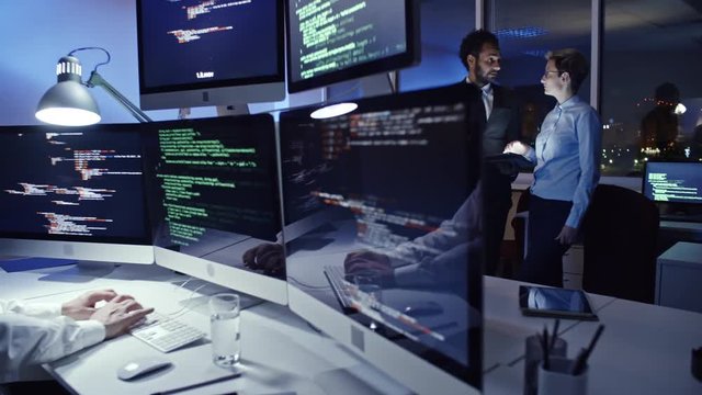 Experienced male cyber security professional working on multi-screen computer in office at night and talking on mobile phone while businesswoman and businessman discussing something on tablet