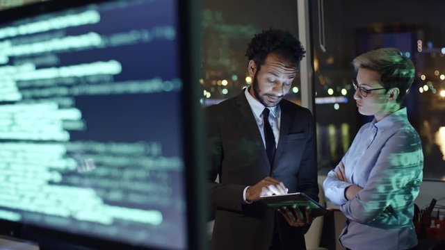 Tracking shot of computer monitors with programming codes and two colleagues discussing something on digital tablet while working in office at night
