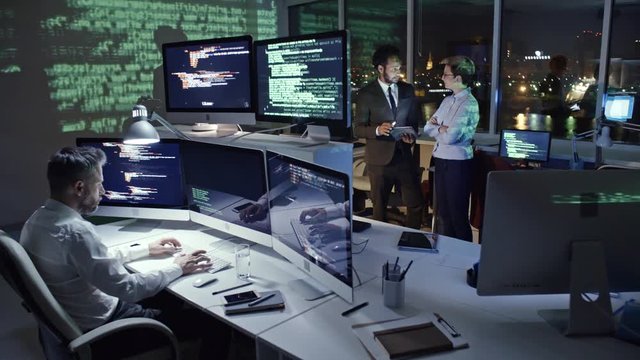 PAN of programmer typing computer code on multiply screen computer in dark office at night while African businessman working on tablet with female colleague; computer data reflecting on the wall