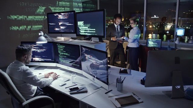 Male programmer typing computer code on multi-screen computer in dark office at night while African businessman working on tablet with female colleague; programming code reflecting on the wall