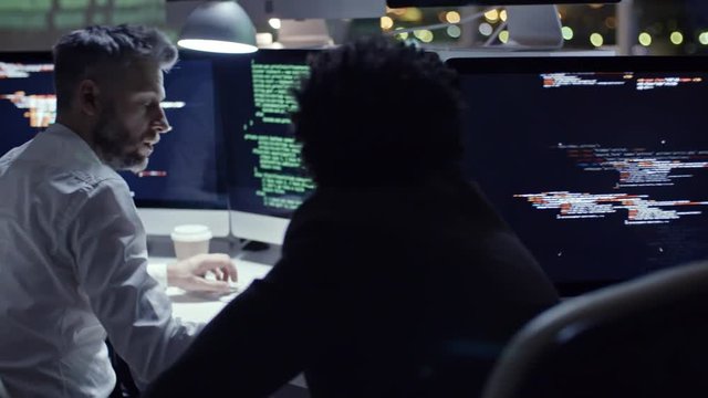 Tracking shot of IT security team working at night in the office: woman typing on computer and male colleagues discussing programming code on computer screen