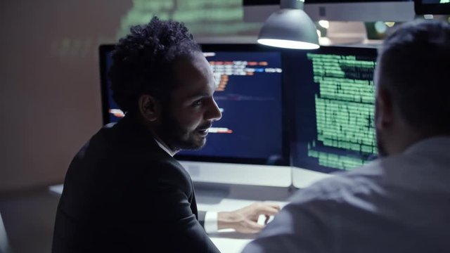 African programmer writing code on computer and talking to his colleague while working in the office at night