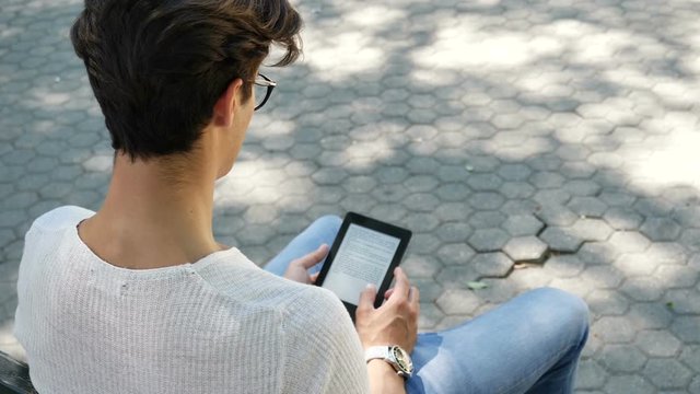 Young attractive man in city park using ebook reader to read a book, sitting on a bench