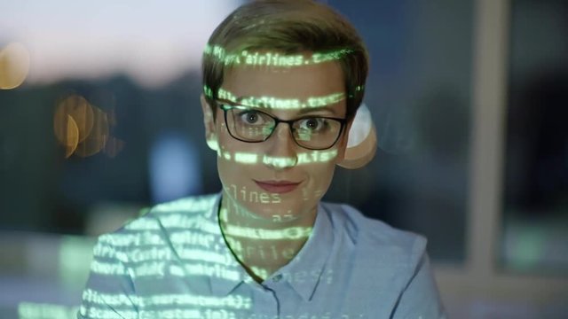 Tilt up of beautiful female programmer typing code on computer in the dark office, looking at camera and smiling. Green source code reflecting on woman
