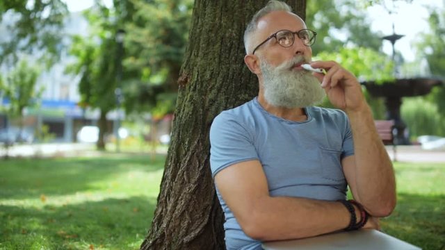 Joyful bearded vaper sitting under tree and smoking