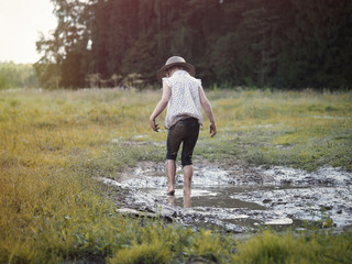 A child playing in a muddy puddle. Dirty girl in a hat and barefoot. Rural road