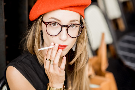 Portrait of a young stylish woman in red beret and eyeglasses smoking a cigarette