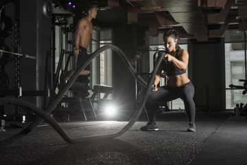 Strong athlete woman wearing a black sports bra and long tights in a dark gym with a muscular male...