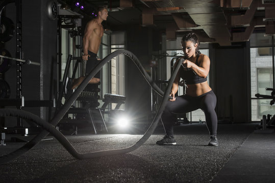 Strong athlete woman wearing a black sports bra and long tights in a dark gym with a muscular male in the background  using  battle ropes for exercise