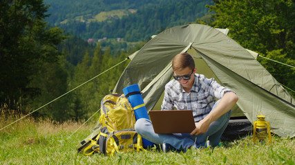 A young male tourist uses a laptop in a camping near a tent. In a picturesque place in the background of the mountains