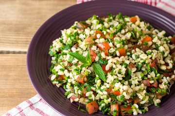 Tabbouleh salad with bulgur, tomatoes, parsley, green onion and mint in plate on wooden table.
