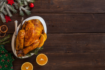 Roasted Christmas chicken on plate and festive new year decoration on dark wooden background. Top view, copy space. Cone, dried oranges, knife, fork furry spruce on a table.
