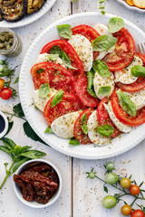 Caprese salad made of sliced fresh tomatoes, mozzarella cheese and basil  served on a white plate on a wooden table, top view.Traditional Italian food