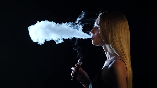 Blonde in a spacious room smokes an electronic cigarette. Black background. Side viwe
