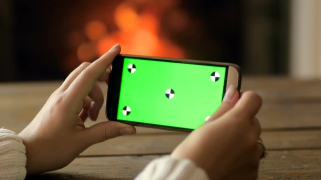 Closeup 4k footage of young woman sitting by the fireplace and holding mobile phone with green chroma key screen