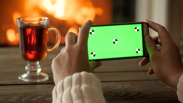 Closeup 4k shot of young woman sitting by the fireplace and holding smartphone with green screen. Good Christmas footage for adding your video on mobile phone chroma key screen