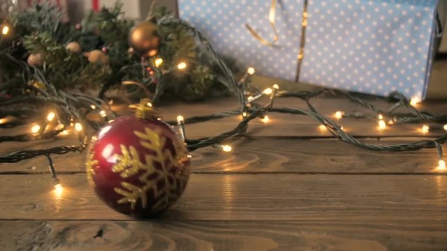 Slow motion footage of red Christmas bauble falling and rolling on table decorated with lights and gift boxes