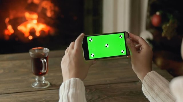 Closeup slow motion footage of woman sitting at fireplace and using smartphone with green screen. Perfect video for Chroma Key