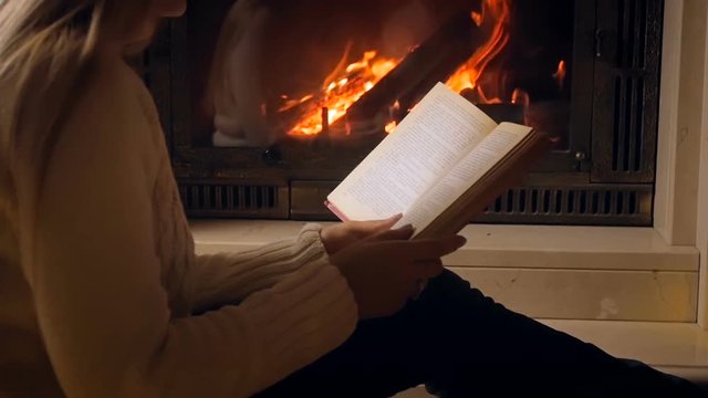 Slow motion video of woma reading book sitting on floor next to burning fireplace in house at night