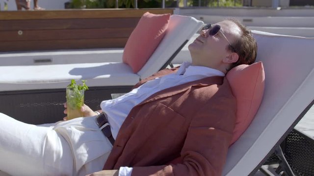 Mixed race Asian/Caucasian man in sunglasses, slacks, white shirt and jacket, relaxes with a cocktail on a sun-lounger at a beautiful resort location. Slow motion hand-held 4K, recorded at 60fps.