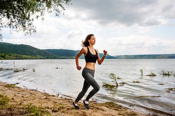 A beautiful sporty woman runing on the shore of a lake in sportswear. Girl is exercising