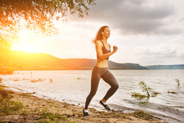 Fototapeta na wymiar A beautiful sporty woman runing on the shore of a lake in sportswear. Girl is exercising