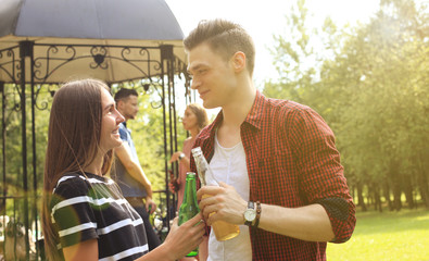 Smiling young couple with beer and looking at each other while two people barbecuing in the background.
