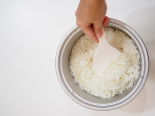 Hand holding big white spoon scooping Thai local traditional boiled white jasmine rice in aluminum cooker, top view, hand vertical