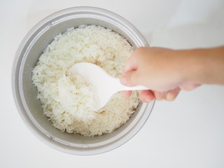 Hand holding big white spoon scooping Thai local traditional boiled white jasmine rice in aluminum cooker, top view horizontal