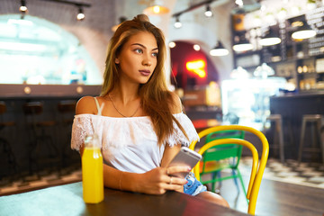 Young, beautiful girl with a lovely smile sitting in a cafe drinking lemonade and using a phone. Excellent summer mood. Background blur

