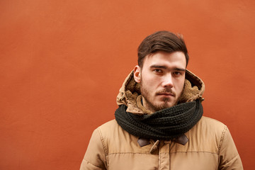 Portrait of serious young bearded man wearing warm winter jacket and black scarf tied around his neck looking at camera, posing isolated at orange blank wall with copy space for your content