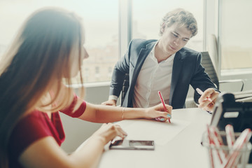 Two business persons (woman in red dress and caucasian handsome man in formal suit) having work meeting in bright office room; woman is explaining something to colleague with drawing on paper sheet