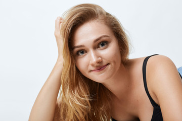 Close up portrait of attractive female with blue eyes, pure skin and light hair, leaning at her hand while lying over white studio background. Beautiful woman feeling relaxation being photographed