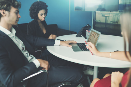 View of meeting room in office settings with group of people having work meeting: caucasian handsome man employer, brazilian young woman probationer and caucasian female co-worker, laptop on the table