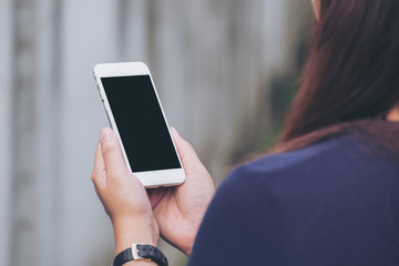 Mockup image of a woman holding and using white smart phone with blank black screen in outdoor and old white concrete wall background