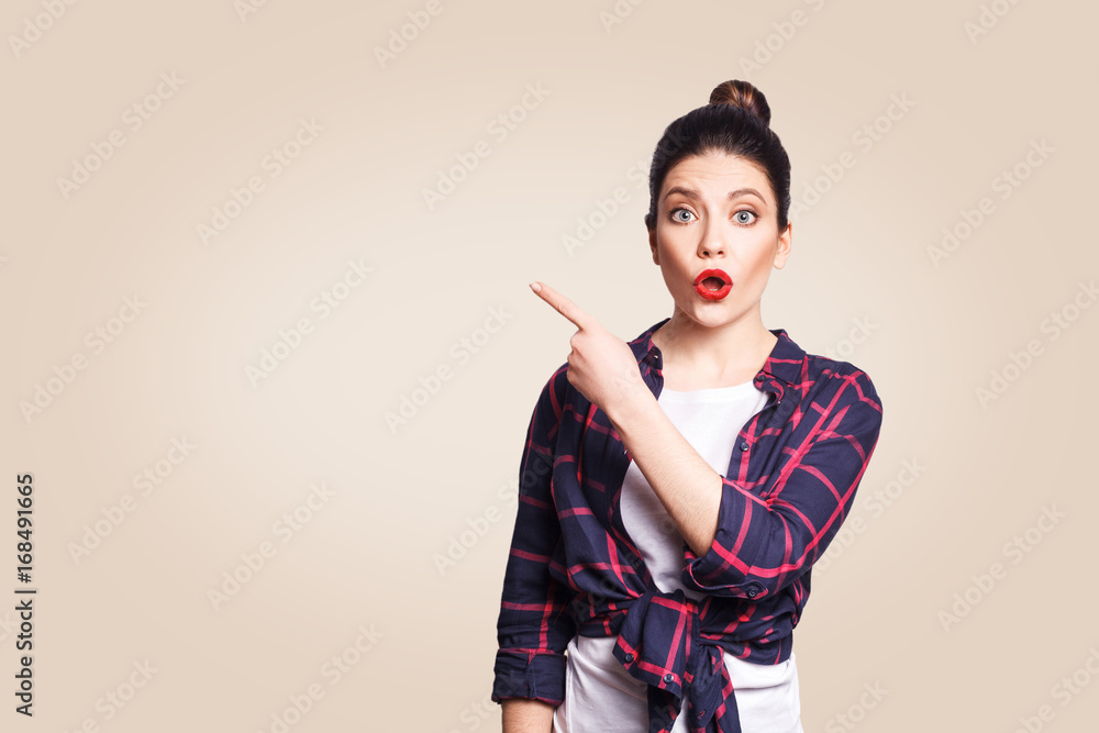 Wall mural young surprised girl with casual style and bun hair pointing her finger sideways, demonstrating some