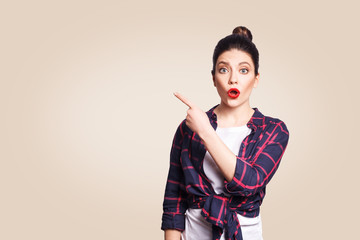 Young surprised girl with casual style and bun hair pointing her finger sideways, demonstrating something on beige blank wall with copy space for your information or promotional content.