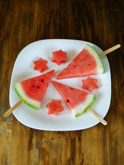 Watermelon. Watermelon pieces formed as popsicles and as stars on a white plate on a wooden background. Unusual watermelon serving