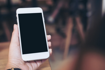 Mockup image of hand holding white mobile phone with blank black screen in modern restaurant