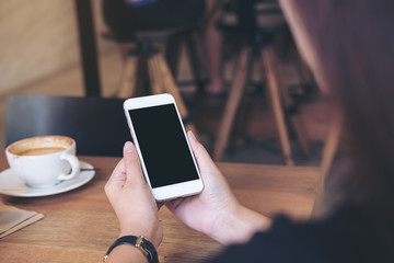 Mockup image of hands holding white mobile phone with blank black screen with coffee cups on wooden table in restaurant