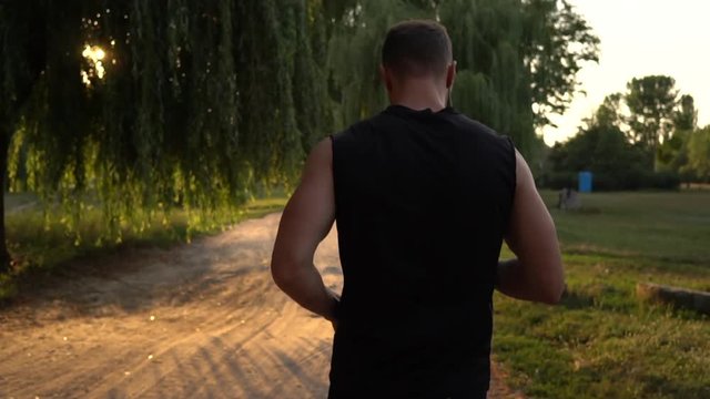 Young male runner runs down the road and listens to music in the park at sunset, slow motion