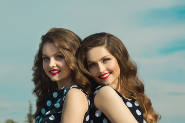 Two beautiful sisters twin girls in identical dresses, with makeup and hairdo on blue sky background