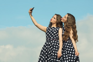 Two beautiful sisters twin girls in identical dresses, with makeup and hairstyle being photographed on the phone against the blue sky