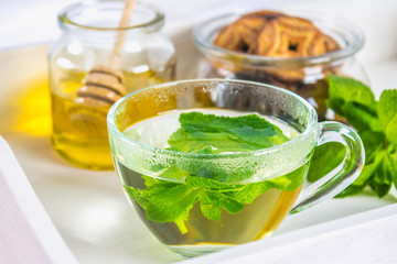 Mint tea in a cup, a jar of honey, a cookie in a jar, on a white tray in bed.