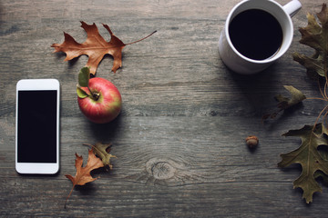 Autumn season still life with red apple, mobile phone, black coffee cup and fall leaves over rustic wooden background