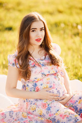 Fototapeta premium Pregnant girl close-up looks into the camera and holds her stomach with her hands. Concept pregnancy, motherhood, expectation of the child. Sunset.