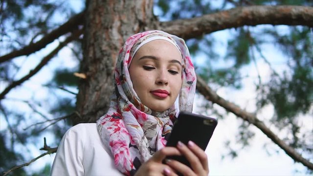 A young Muslim woman in a traditional headscarf holding a smartphone in his hands, a lady communicating with friends on her mobile phone in the park, she sits outside near a tall tree